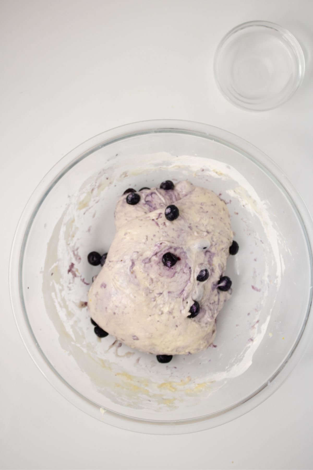 A glass bowl containing dough with blueberries mixed in, placed on a white surface. An empty glass bowl is visible in the upper right corner.