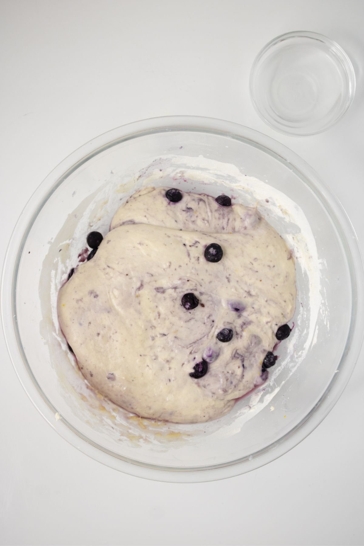A clear glass bowl containing risen blueberry-studded dough sits on a white surface beside an empty small glass bowl.