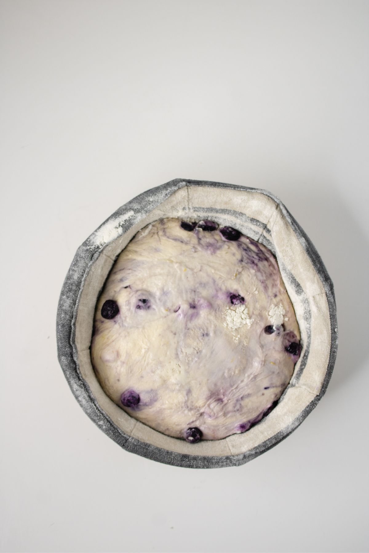 A round dough with visible blueberries rests in a floured proofing basket on a white surface.