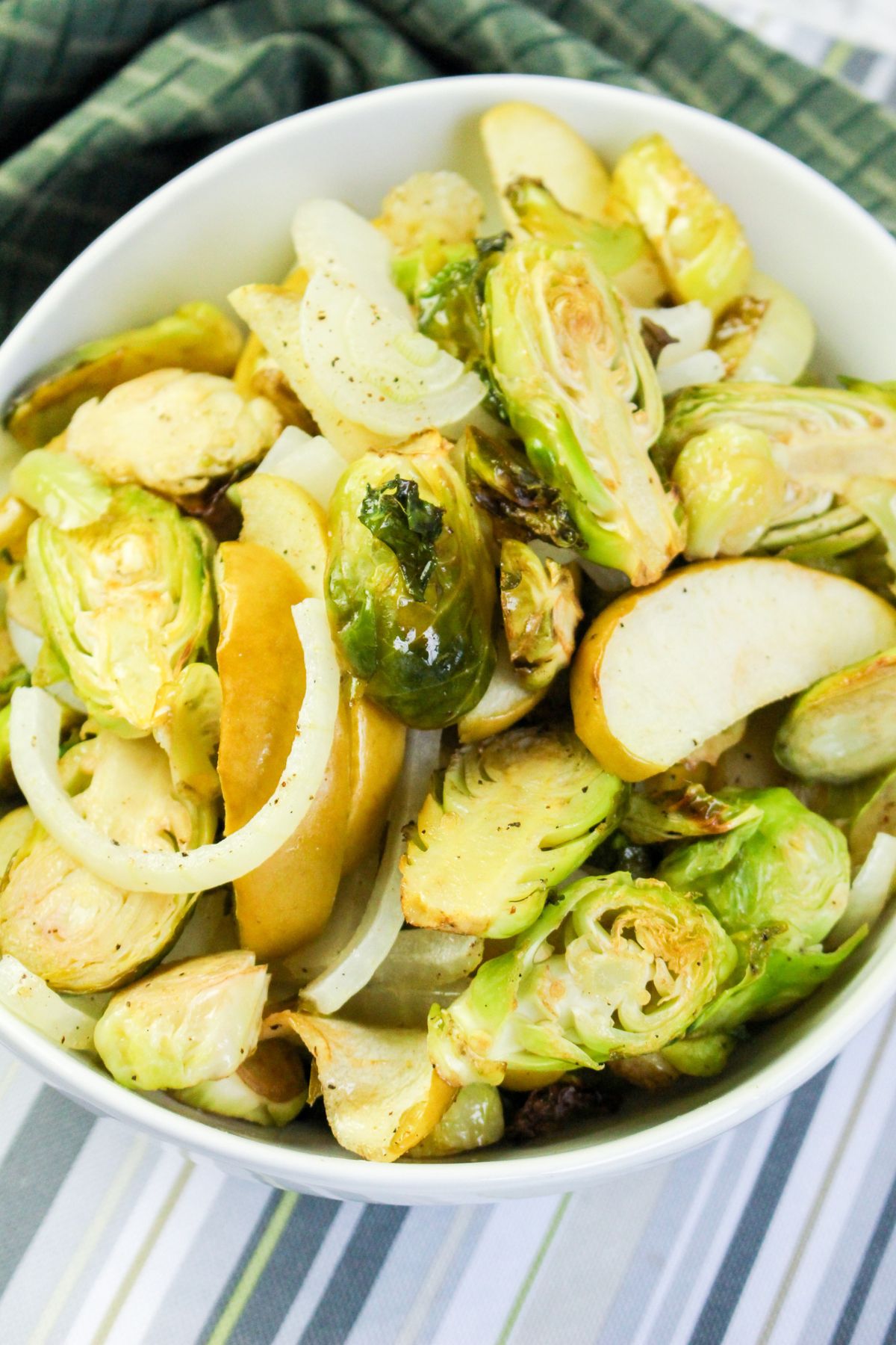A white bowl filled with roasted Brussels sprouts, sliced onions, and apple wedges, set on a striped surface with a green cloth in the background.