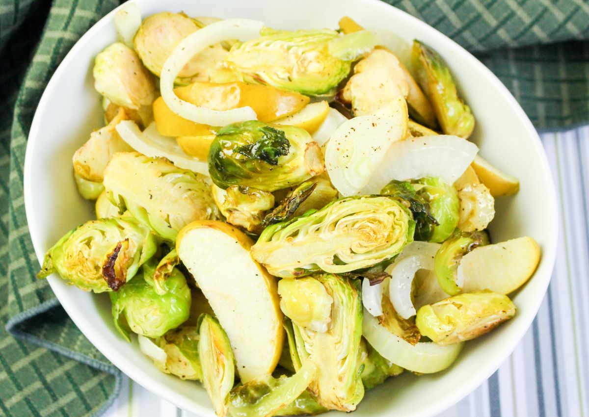 A white bowl filled with roasted Brussels sprouts, sliced onions, and apple wedges on a striped surface with a green cloth nearby.