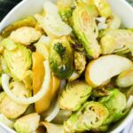 A bowl of roasted Brussels sprouts, sliced onions, and apple wedges, seasoned with black pepper.