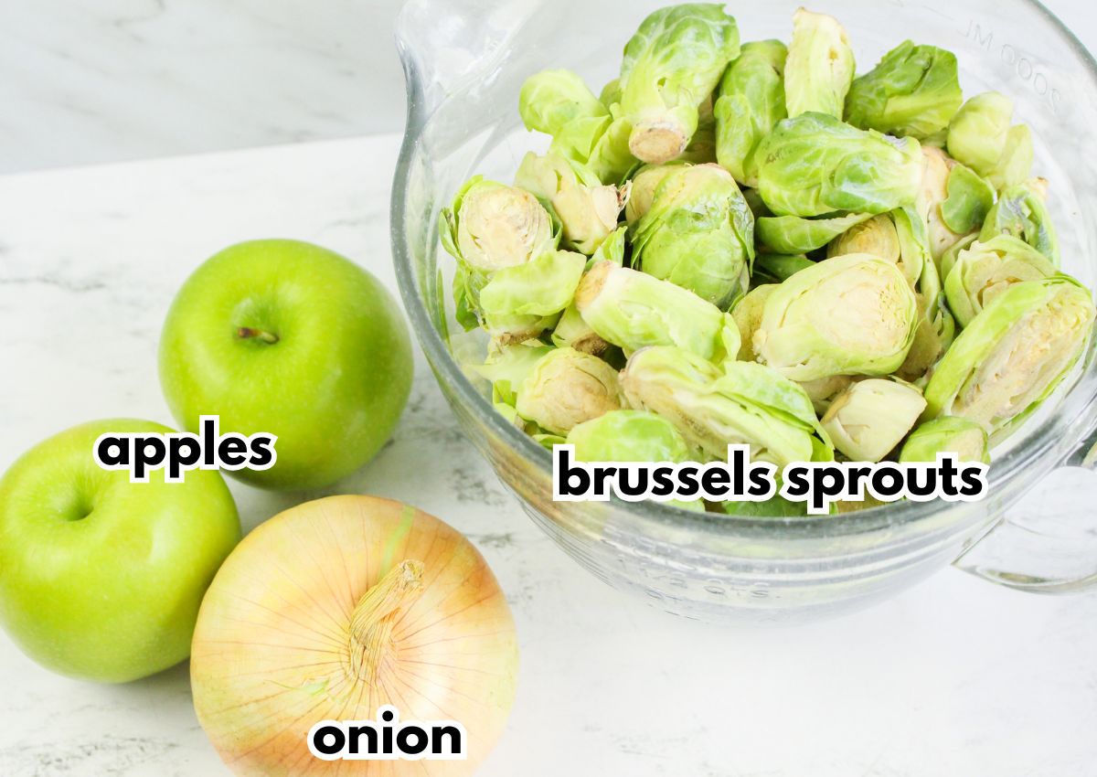 A glass bowl of halved brussels sprouts next to two green apples and a yellow onion on a white surface, each labeled.
