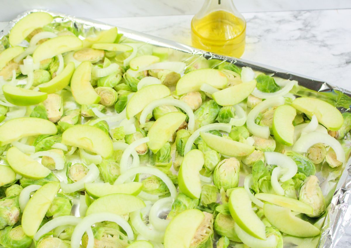 A baking sheet lined with foil holds sliced Brussels sprouts, green apple wedges, and onion rings, with a bottle of oil in the background.