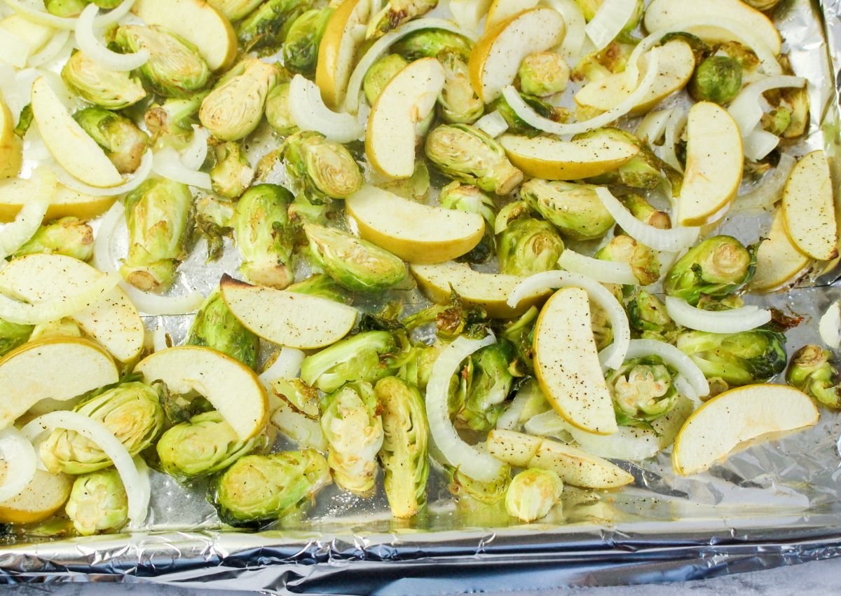 Sliced Brussels sprouts, onions, and apples are spread on a foil-lined baking sheet, seasoned and ready for roasting.