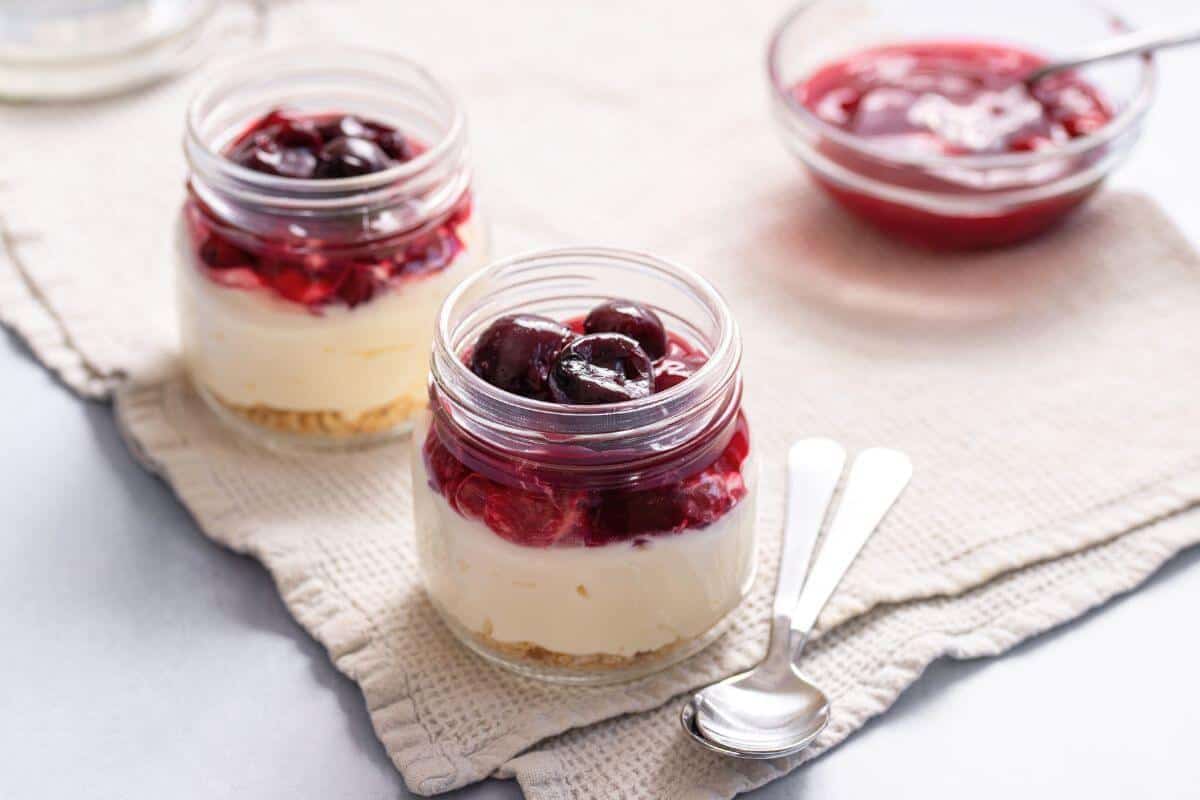 Two small glass jars filled with layered cheesecake and cherry topping sit on a cloth, with a bowl of cherry sauce and two spoons beside them.