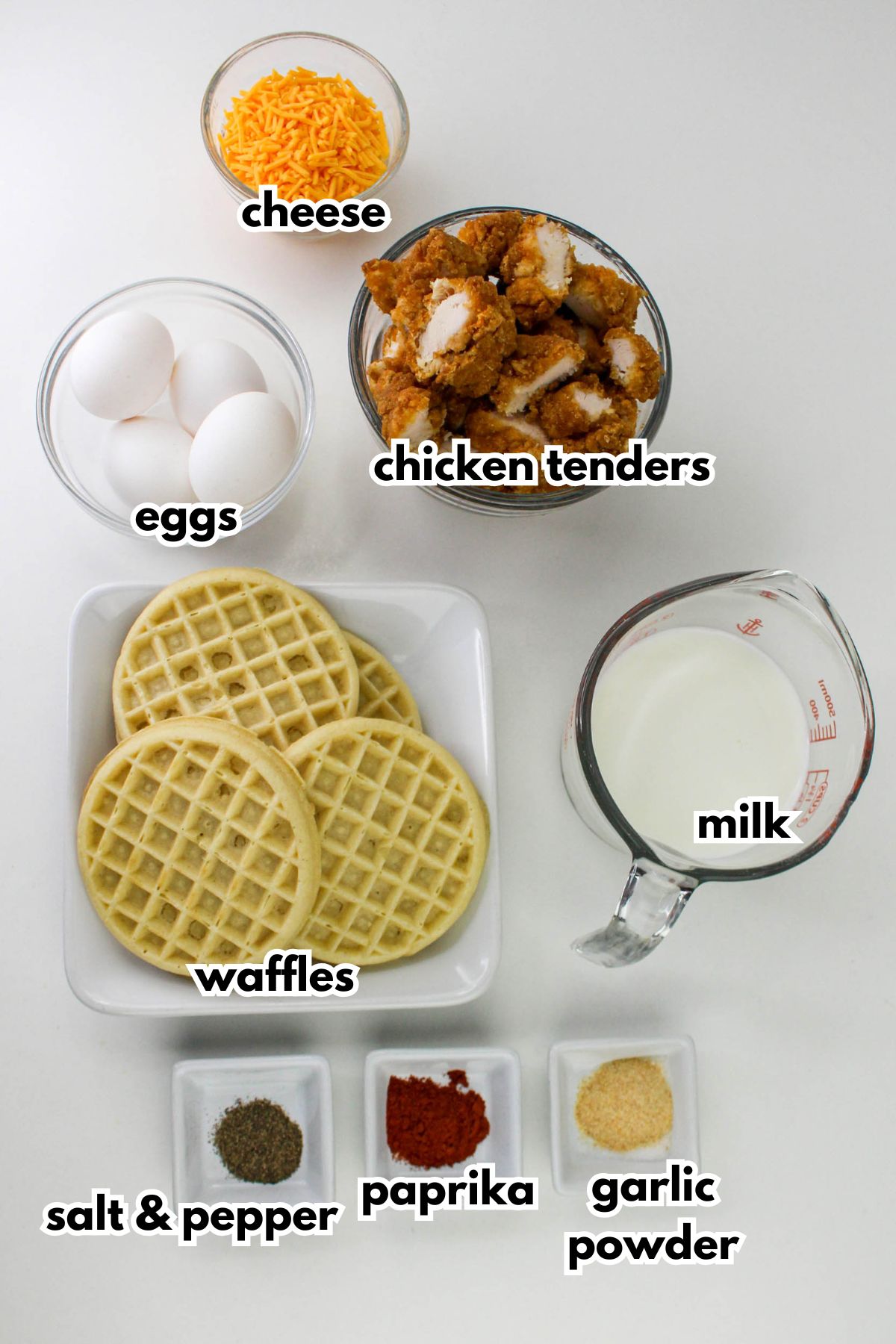 A flat lay of ingredients for chicken and waffles: cheese, eggs, chicken tenders, waffles, milk, salt and pepper, paprika, and garlic powder in separate containers.