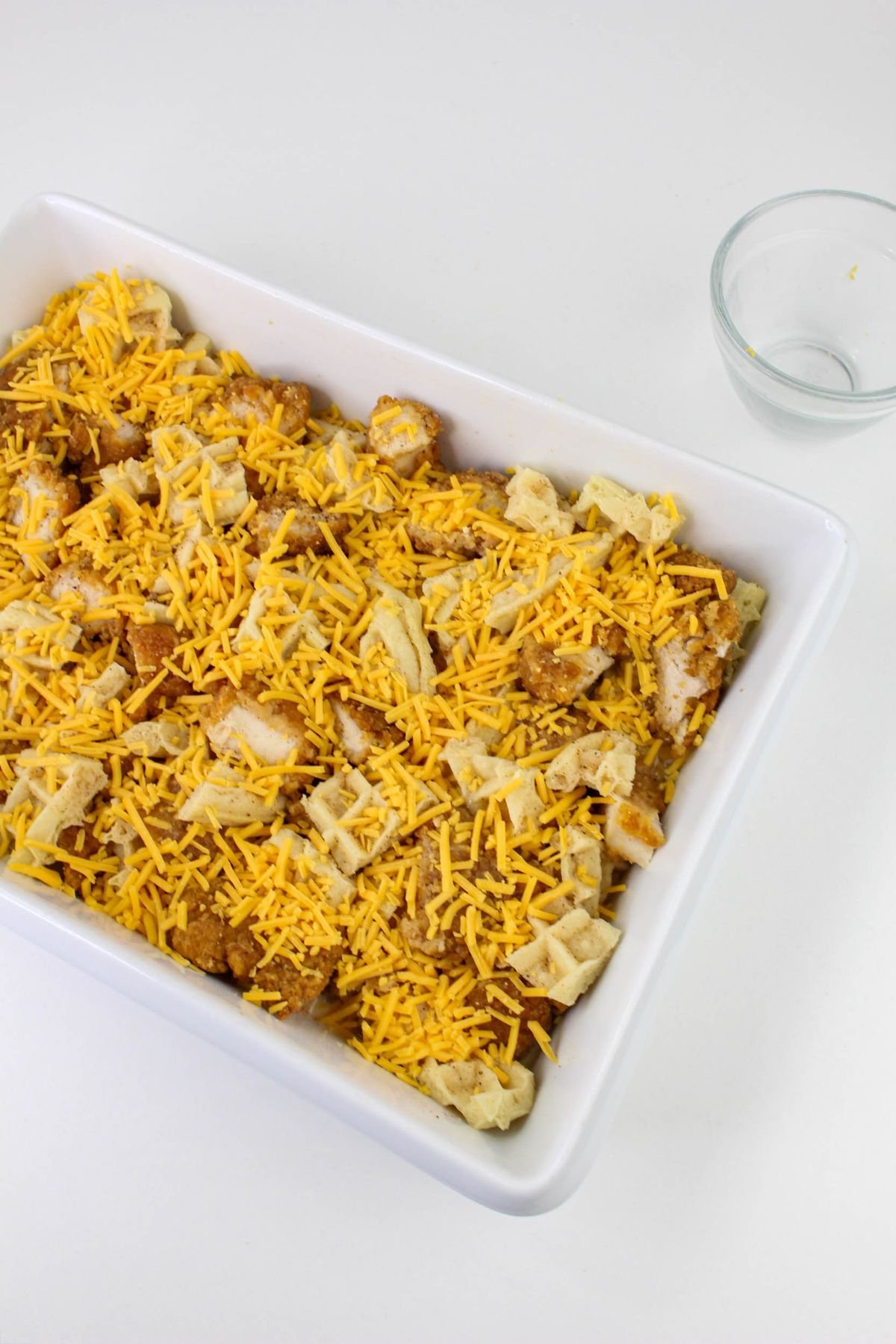 A white baking dish filled with a mixture of waffle pieces, breaded chicken, and shredded cheddar cheese sits next to an empty glass bowl on a white surface.
