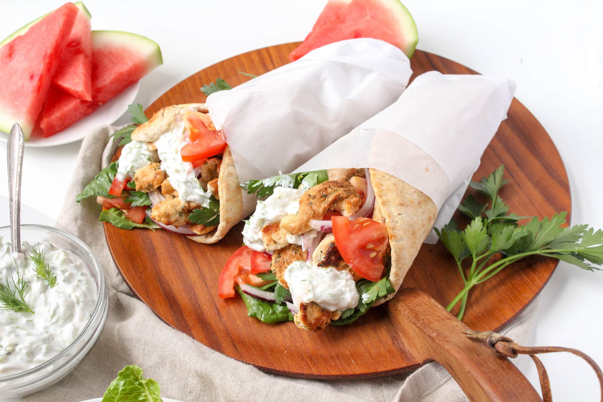 A plate with a two chicken gyros wrapped in parchment paper, with a small bowl of tzatziki sauce beside it.