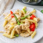 A white plate with penne pasta, shredded chicken, asparagus spears, and red bell pepper pieces, garnished with herbs.