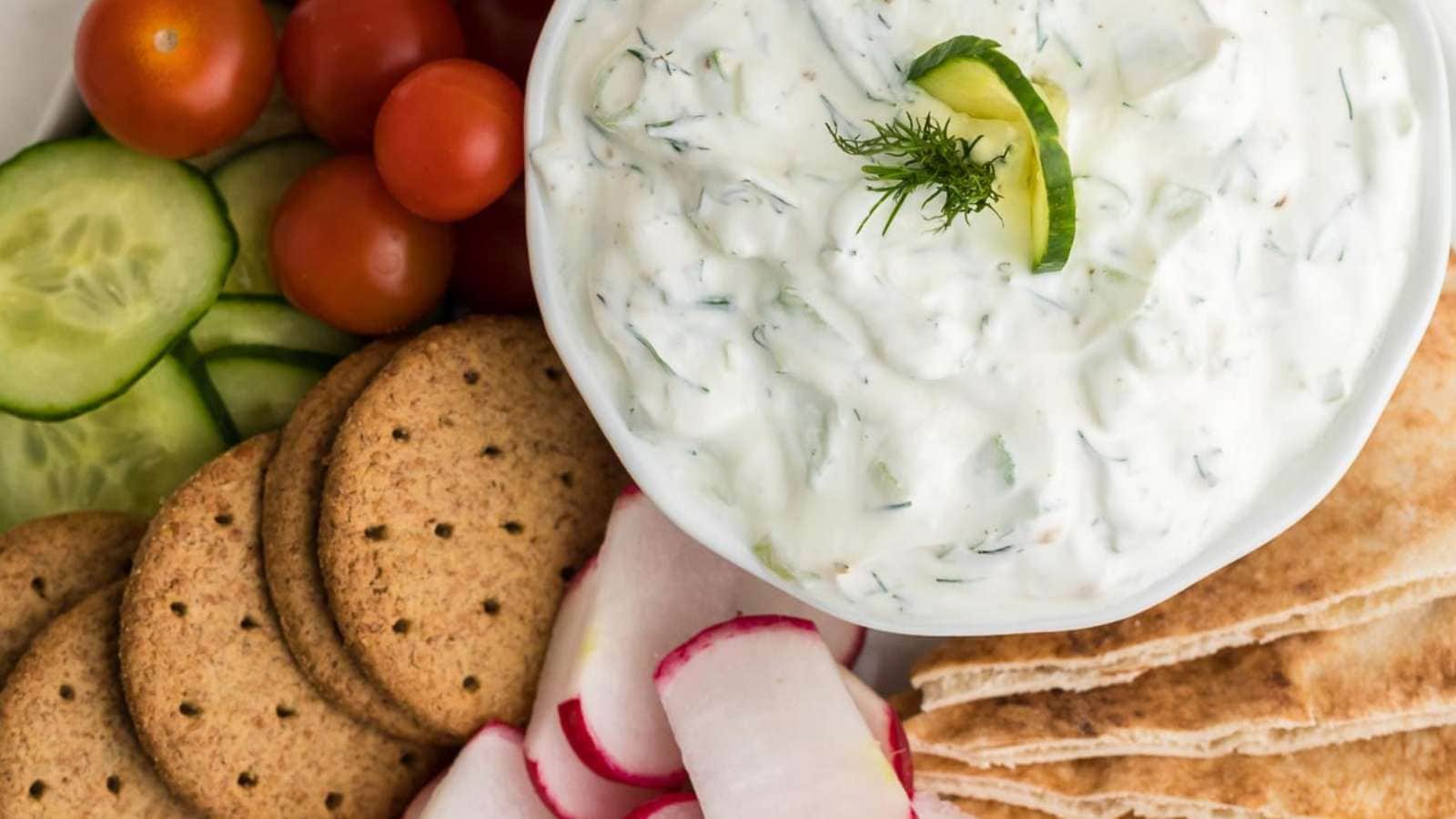 A bowl of tzatziki sauce surrounded by sliced cucumbers, cherry tomatoes, crackers, radishes, and pita bread.