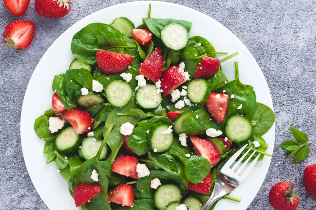 A white plate with a salad made of spinach, sliced cucumbers, strawberries, and crumbled feta cheese, with a fork on the side.