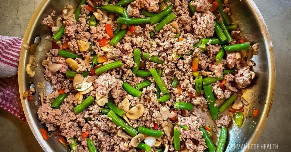 A skillet filled with cooked ground beef, sliced mushrooms, green beans, and diced red bell peppers.