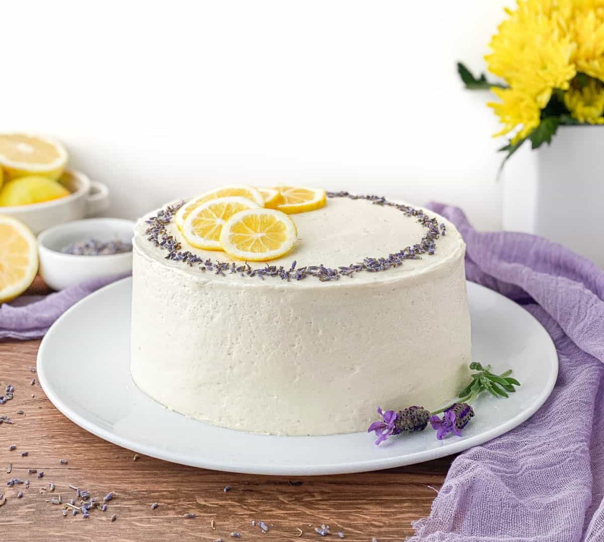 A round frosted cake garnished with lemon slices and purple flowers sits on a white plate, with lemons and yellow flowers in the background.