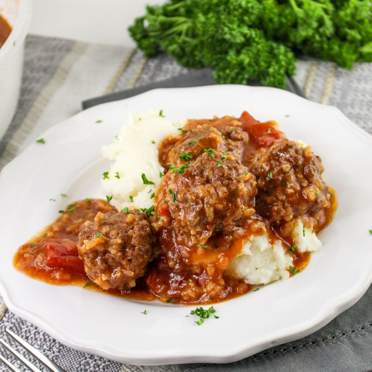 Three beef meatballs in tomato gravy served over mashed potatoes, garnished with chopped parsley, on a white plate.