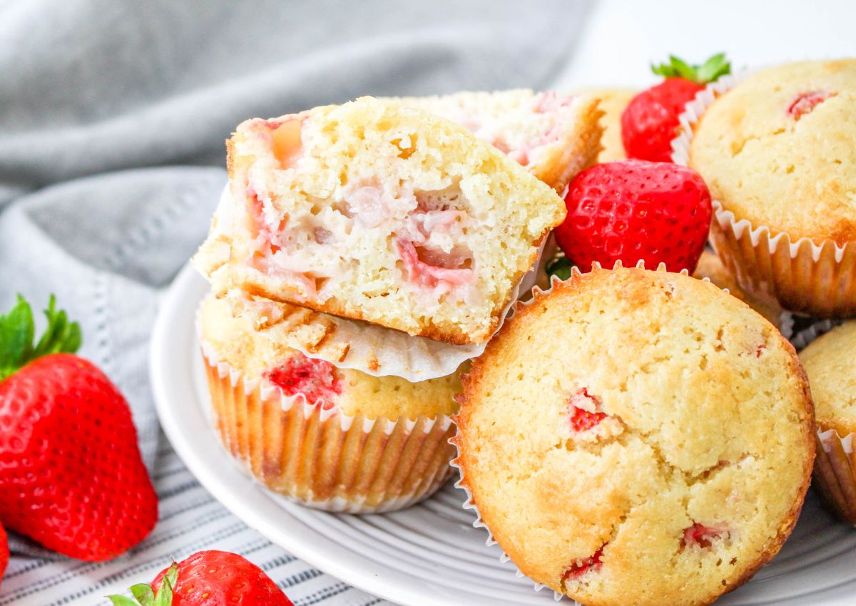 A plate of strawberry muffins, some whole and one cut in half to show the inside, with fresh strawberries around the plate.
