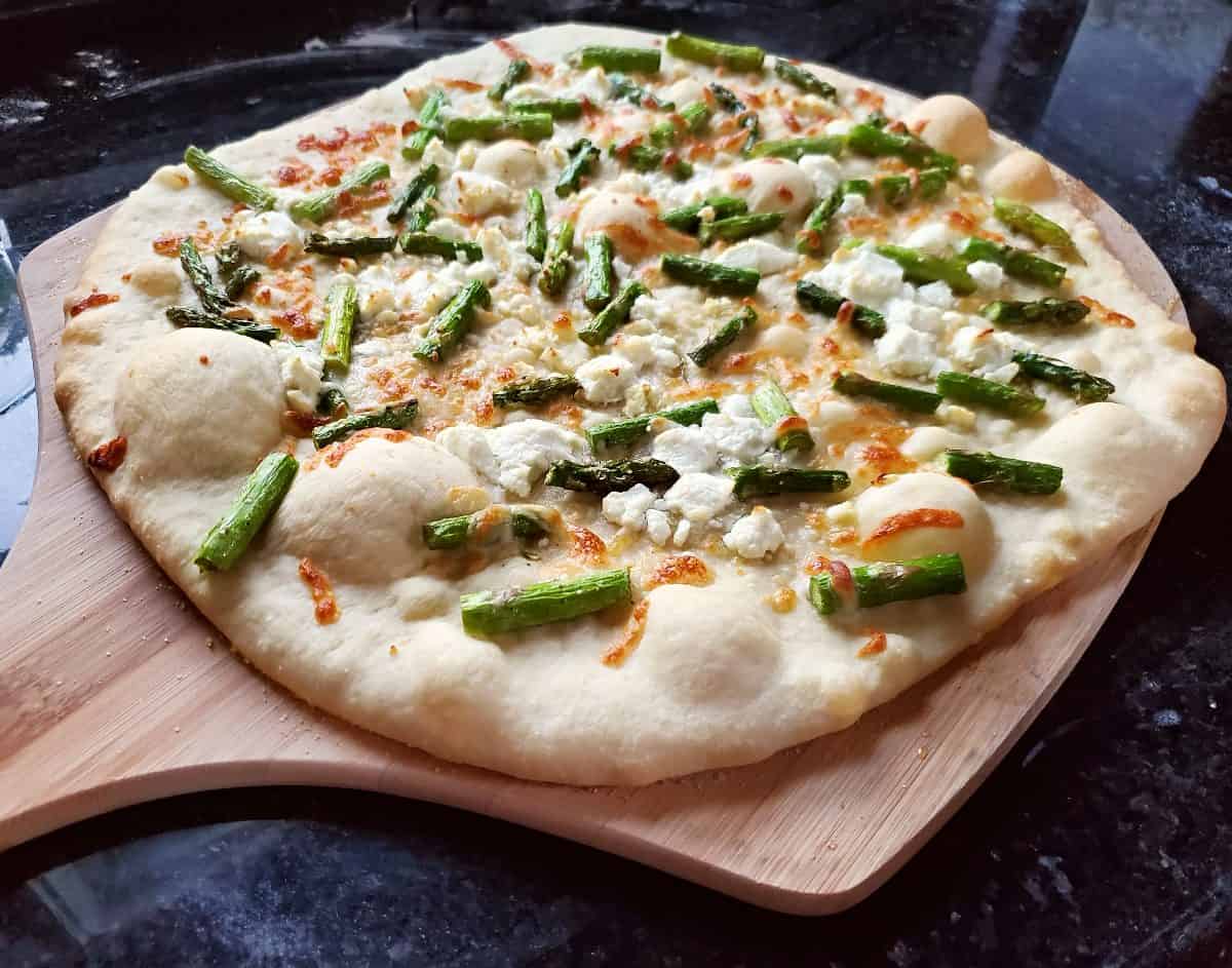 Image shows a close up of an Asparagus and Goat Cheese Pizza on a pizza peel sitting on a black counter.