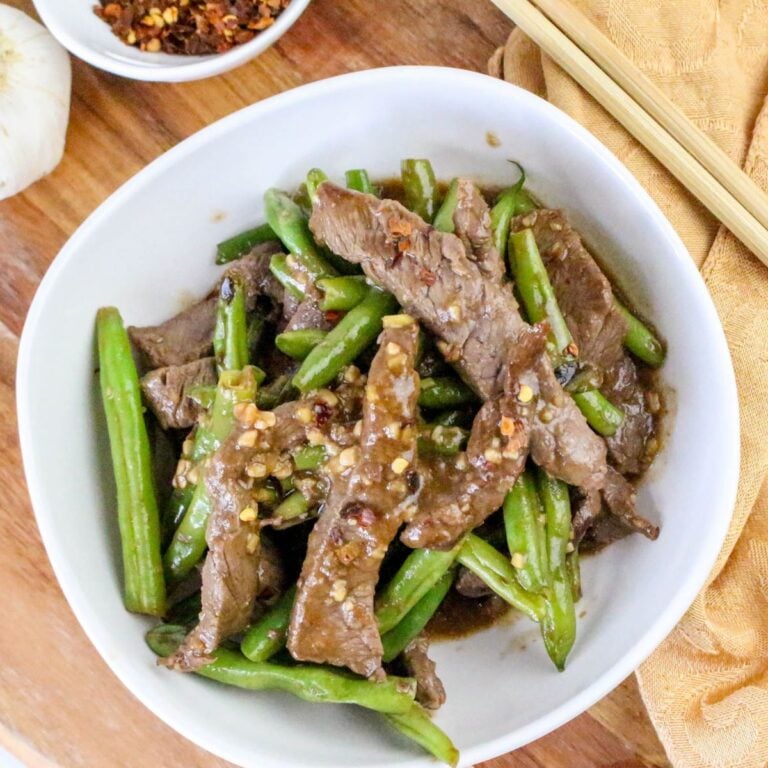 A white bowl filled with stir-fried beef strips and green beans, garnished with crushed red pepper flakes, on a wooden surface with chopsticks nearby.