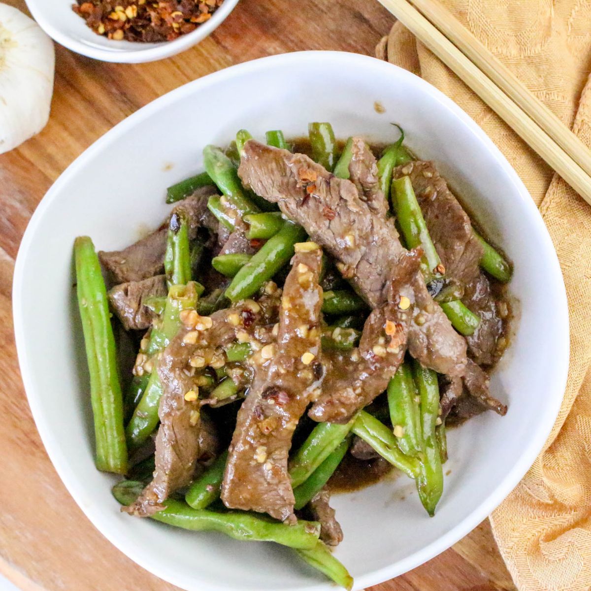 A white bowl filled with stir-fried beef strips and green beans, garnished with crushed red pepper flakes, on a wooden surface with chopsticks nearby.