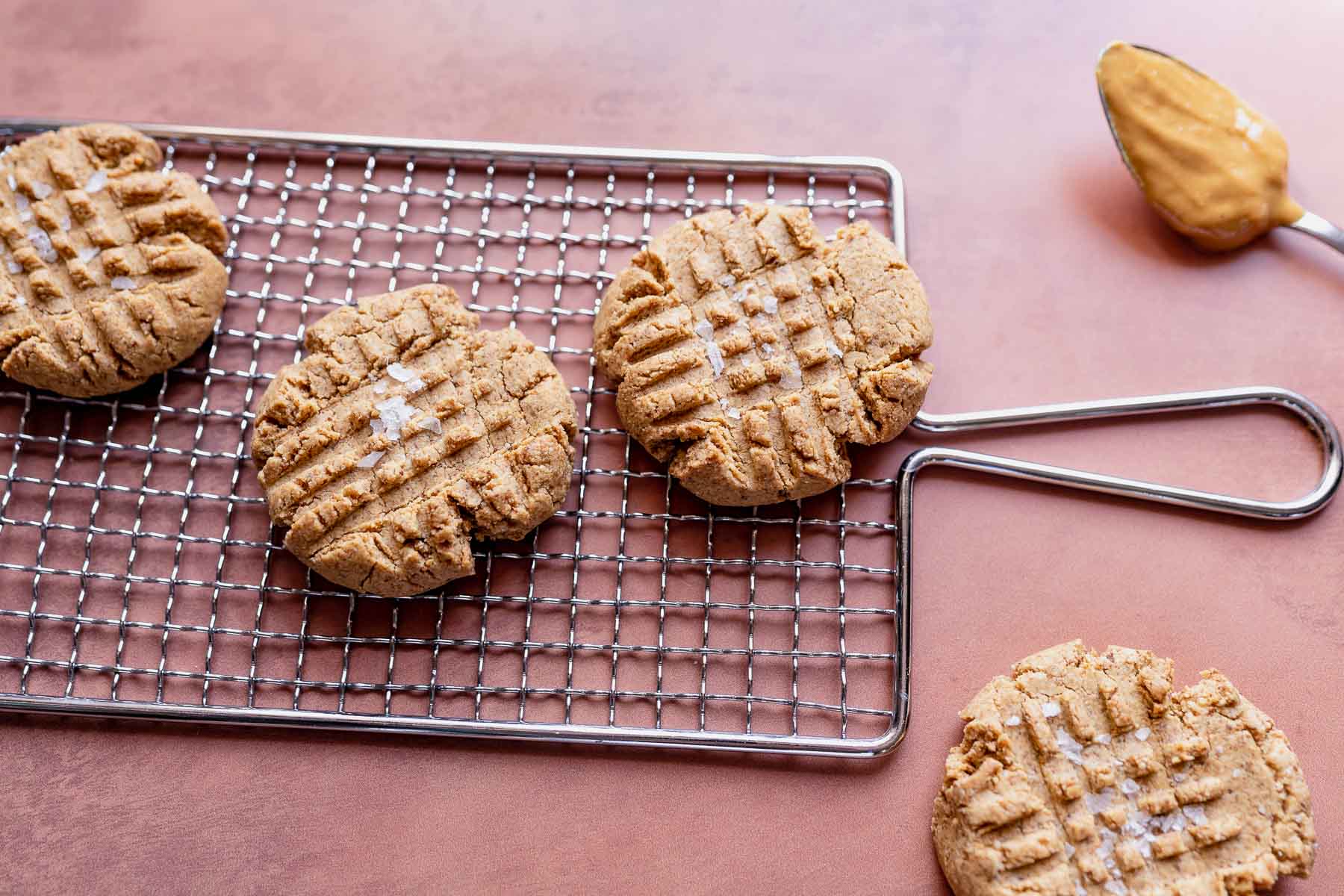 Four peanut butter cookies with fork marks rest on a wire cooling rack, with one cookie on the table and a spoonful of peanut butter nearby.