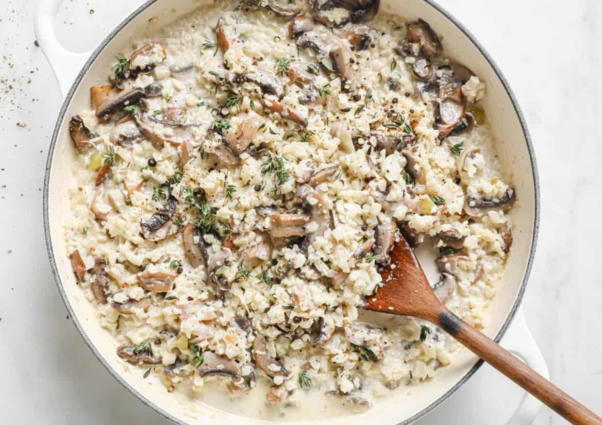 A creamy mushroom risotto with herbs in a white pan, stirred with a wooden spoon.