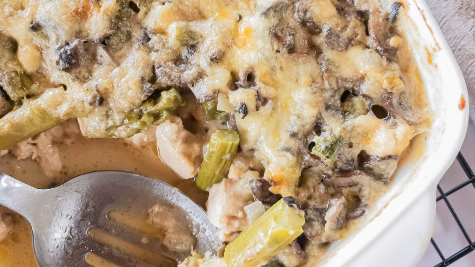 Close-up of a baked casserole with melted cheese, mushrooms, asparagus, and pieces of chicken, with a metal serving spoon in the dish.