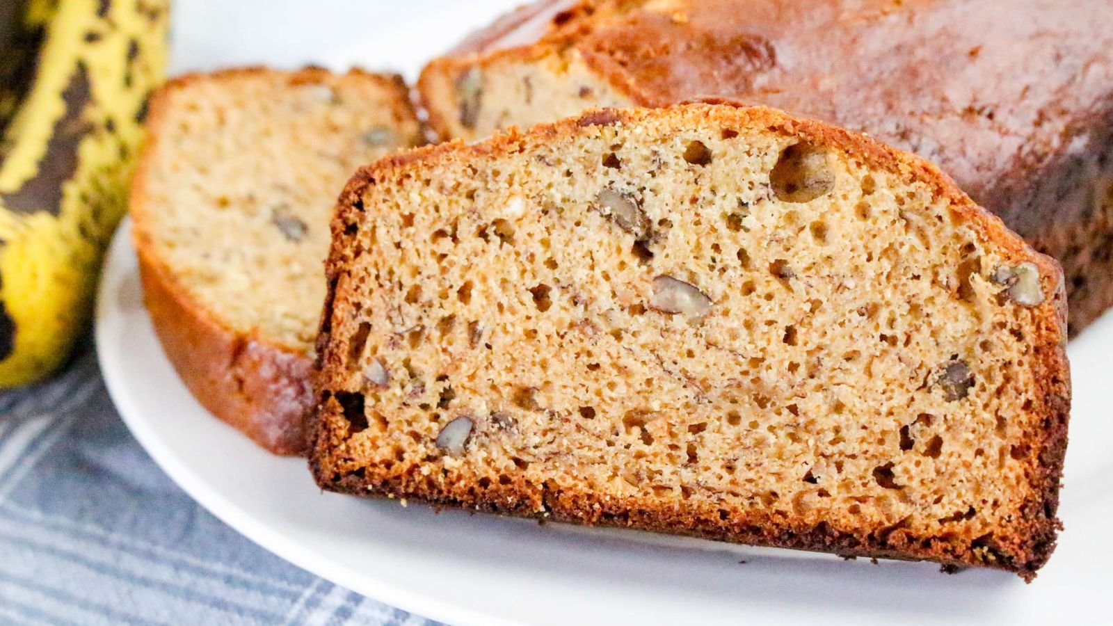 A close-up of sliced banana bread with visible walnuts on a white plate, with overripe bananas in the background.