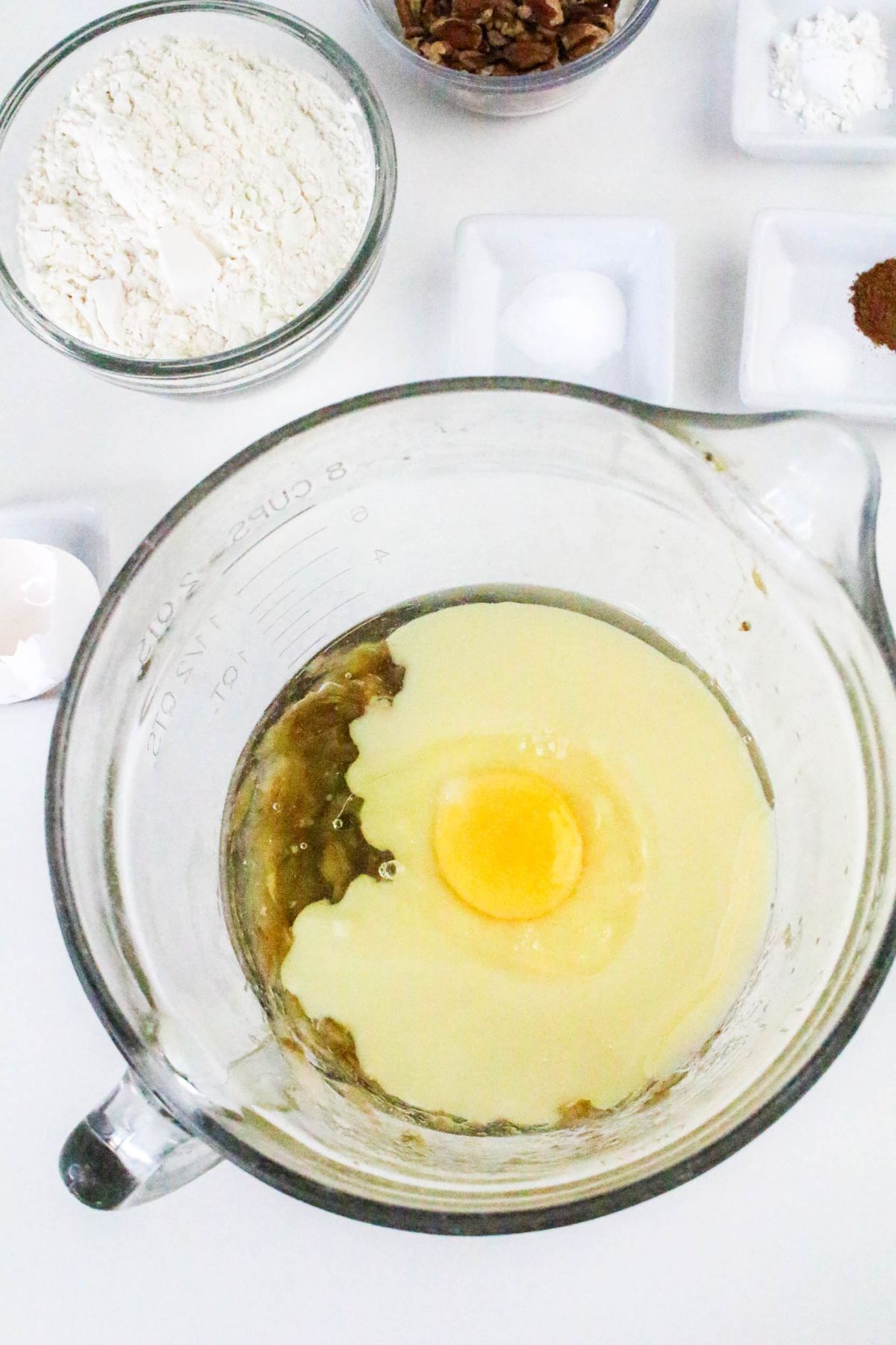 A glass mixing bowl with egg and wet ingredients, surrounded by small bowls of flour, pecans, salt, and spices on a white surface.