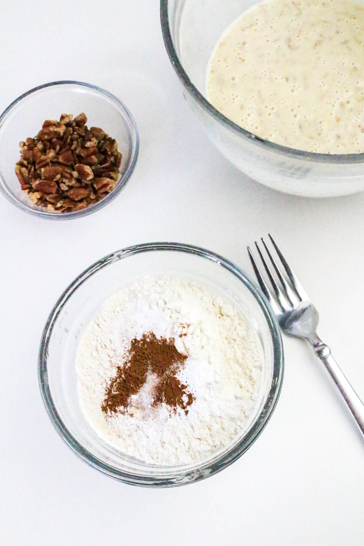 Three glass bowls containing chopped pecans, flour with spices, and a wet batter, with a metal fork placed beside them on a white surface.