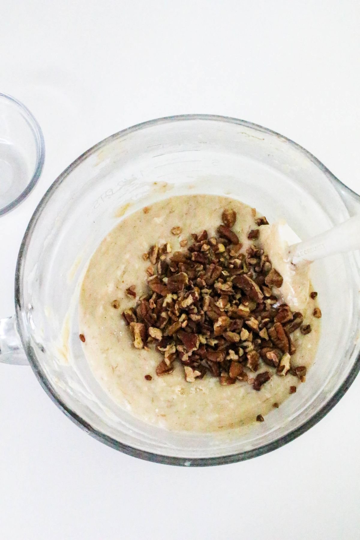A glass mixing bowl containing batter with chopped nuts added on top, and a spatula resting inside the bowl.
