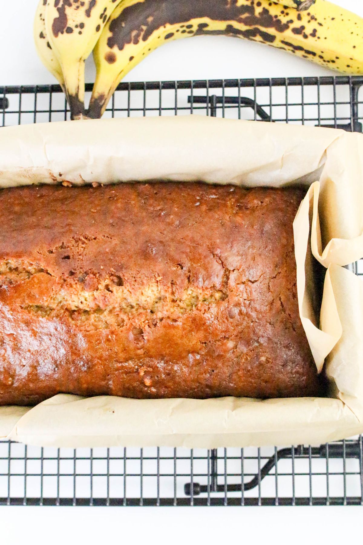 A loaf of banana bread in a parchment-lined pan is cooling on a wire rack, with two ripe bananas in the background.