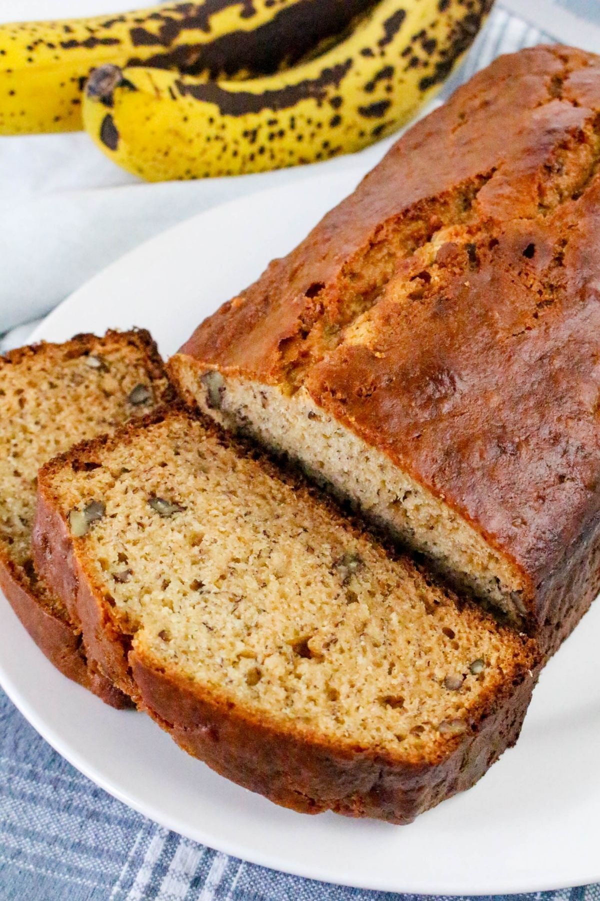 A loaf of banana bread with two slices cut, displayed on a white plate. Ripe bananas with brown spots are in the background.