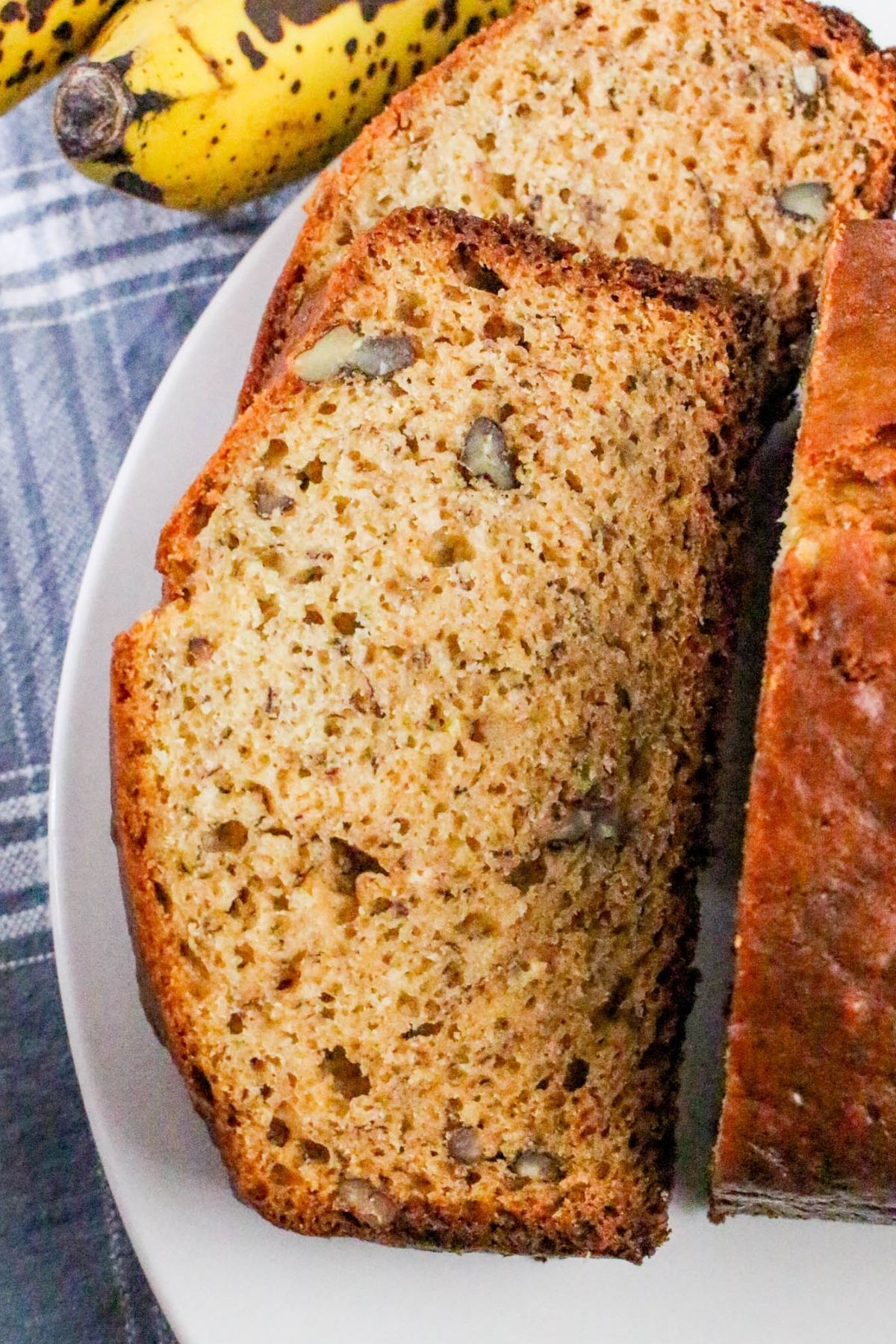 Two slices of banana bread with visible nuts rest on a white plate, with ripe bananas in the background and a blue plaid cloth partially visible.