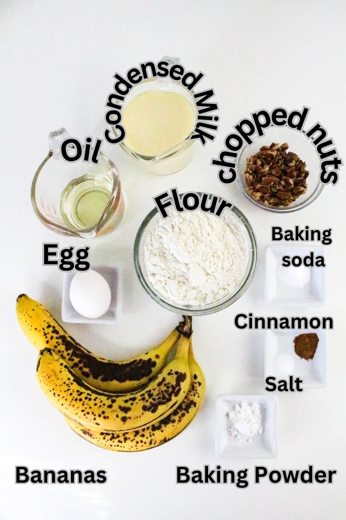 Top-down view of baking ingredients labeled: oil, condensed milk, chopped nuts, flour, egg, baking soda, cinnamon, salt, bananas, and baking powder on a white surface.