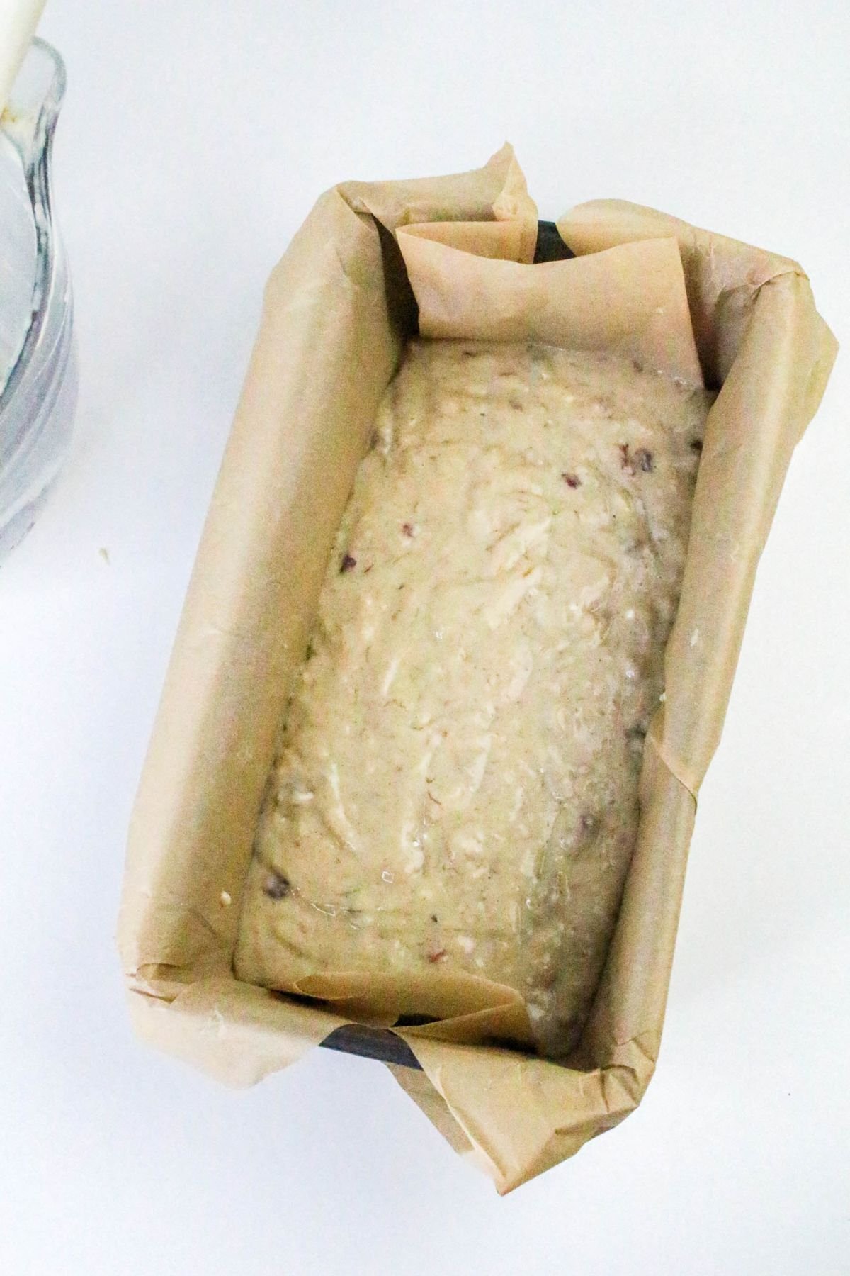 A loaf pan lined with parchment paper filled with unbaked bread or cake batter sits on a white surface.