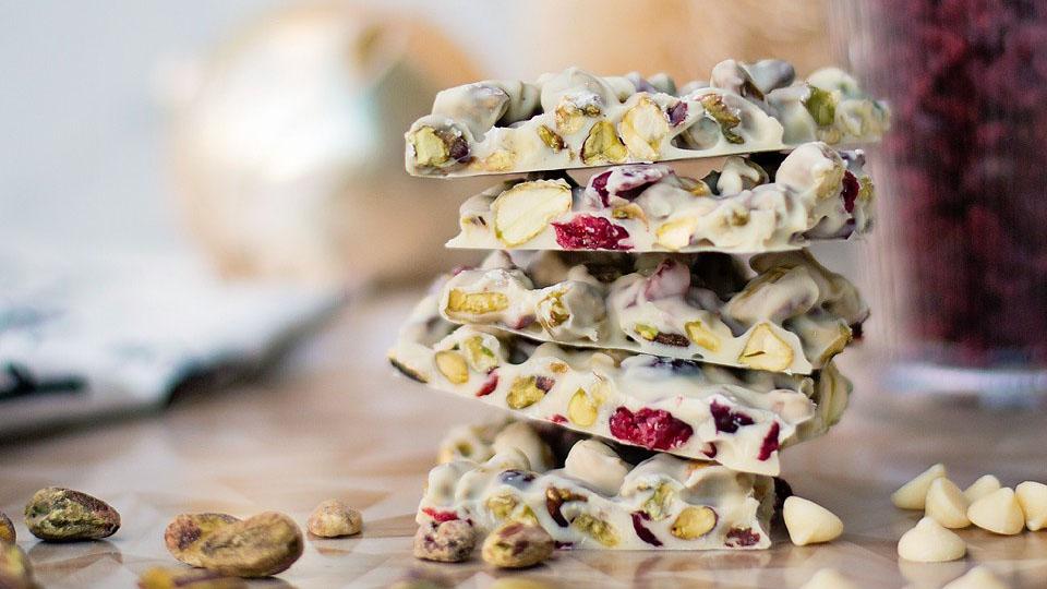 Five stacked pieces of white chocolate bark with pistachios and dried cranberries, with scattered nuts and chocolate chips around them.