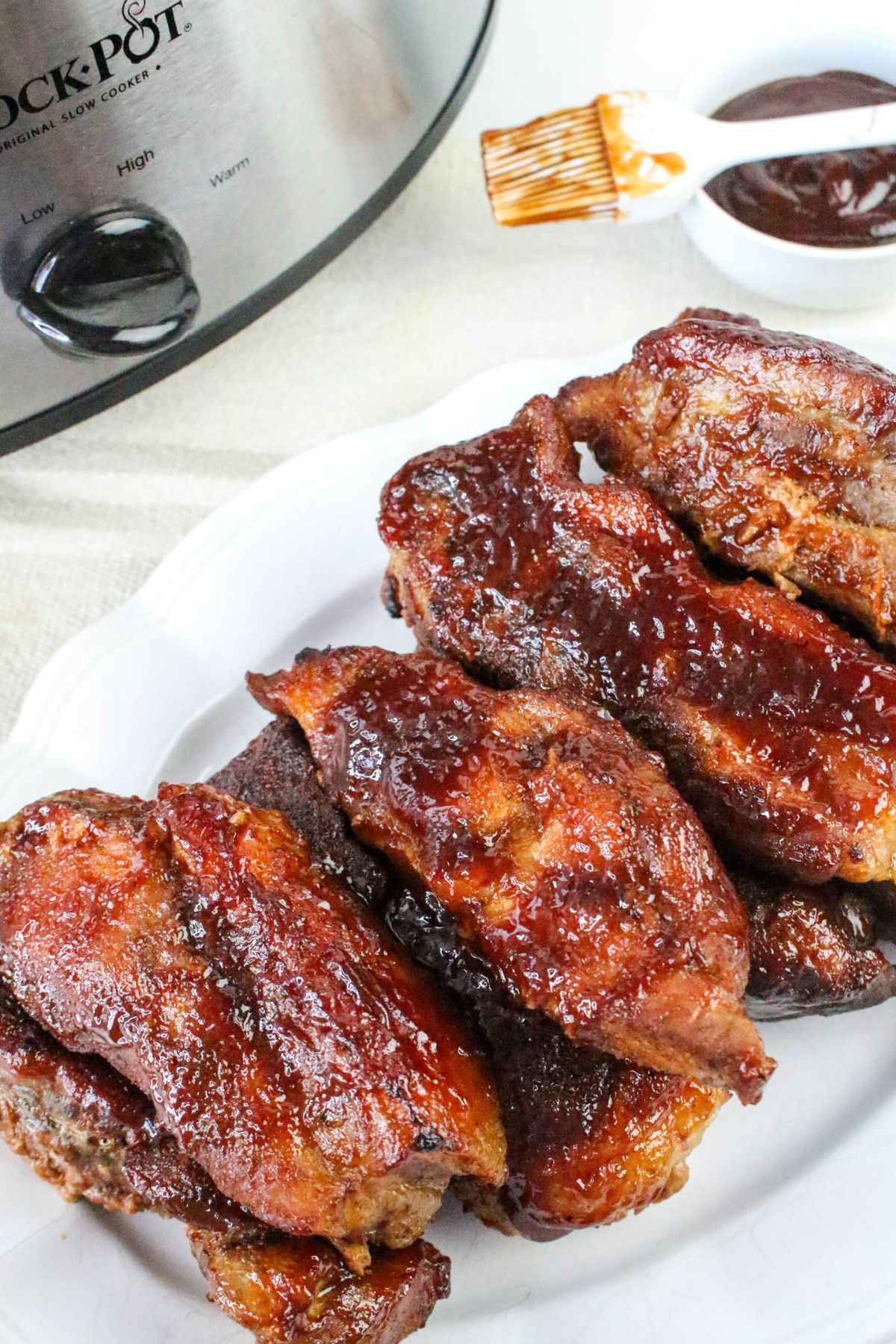A white plate with several pieces of glazed barbecue ribs, a slow cooker, and a small bowl of barbecue sauce with a basting brush in the background.