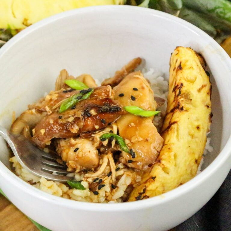 A bowl of rice topped with shredded chicken in sauce, garnished with green onions and black sesame seeds, served with a slice of grilled pineapple.