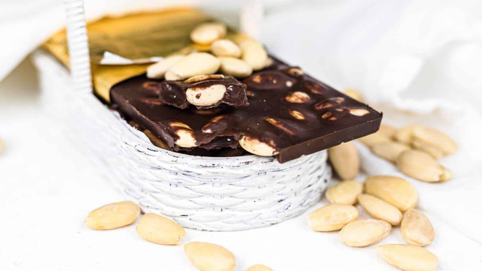 Dark chocolate bar with whole almonds, some broken pieces, and loose almonds displayed in a white basket on a white surface.