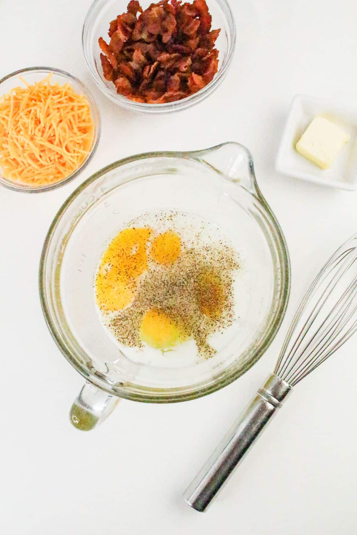 A mixing bowl with eggs, salt, and pepper, surrounded by a whisk, a bowl of shredded cheese, a bowl of chopped bacon, and a small dish of butter on a white surface.