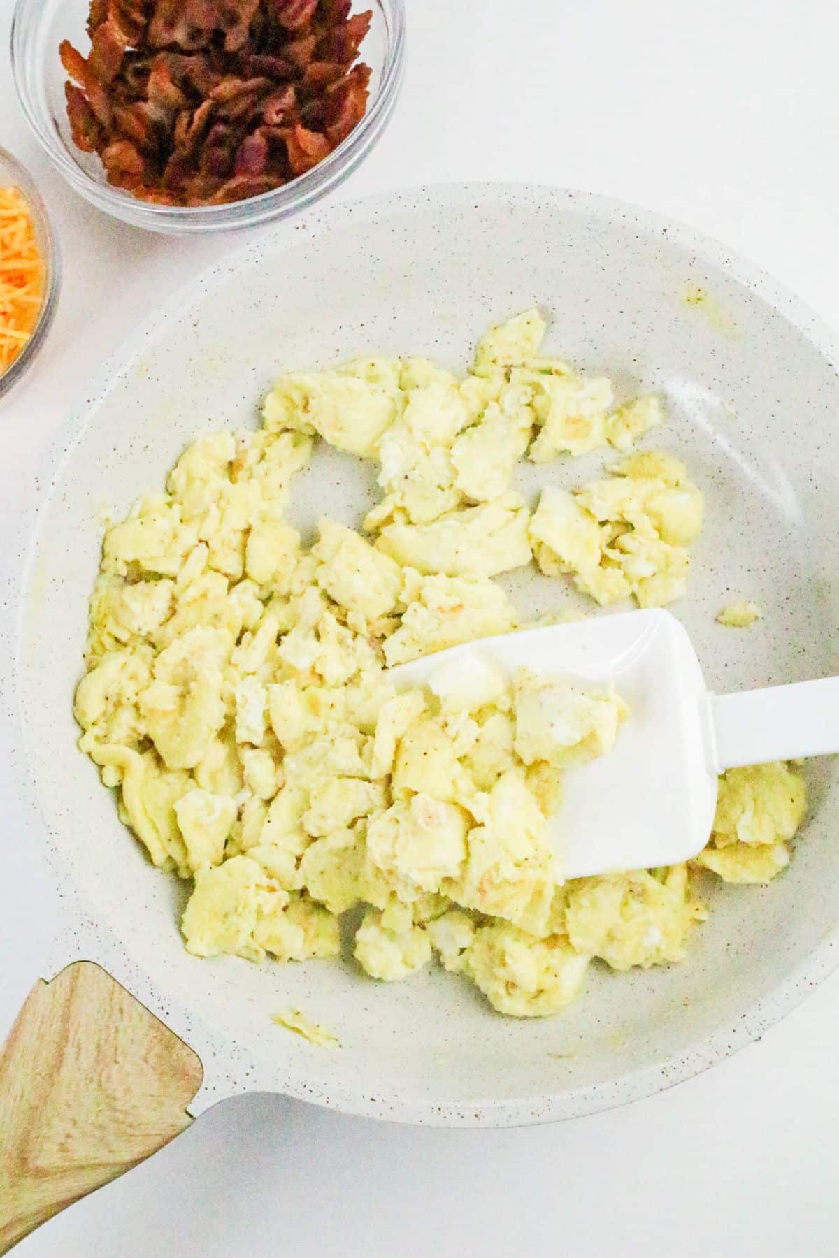 Scrambled eggs being cooked in a white frying pan with a white spatula; bowls of shredded cheese and cooked bacon are nearby.