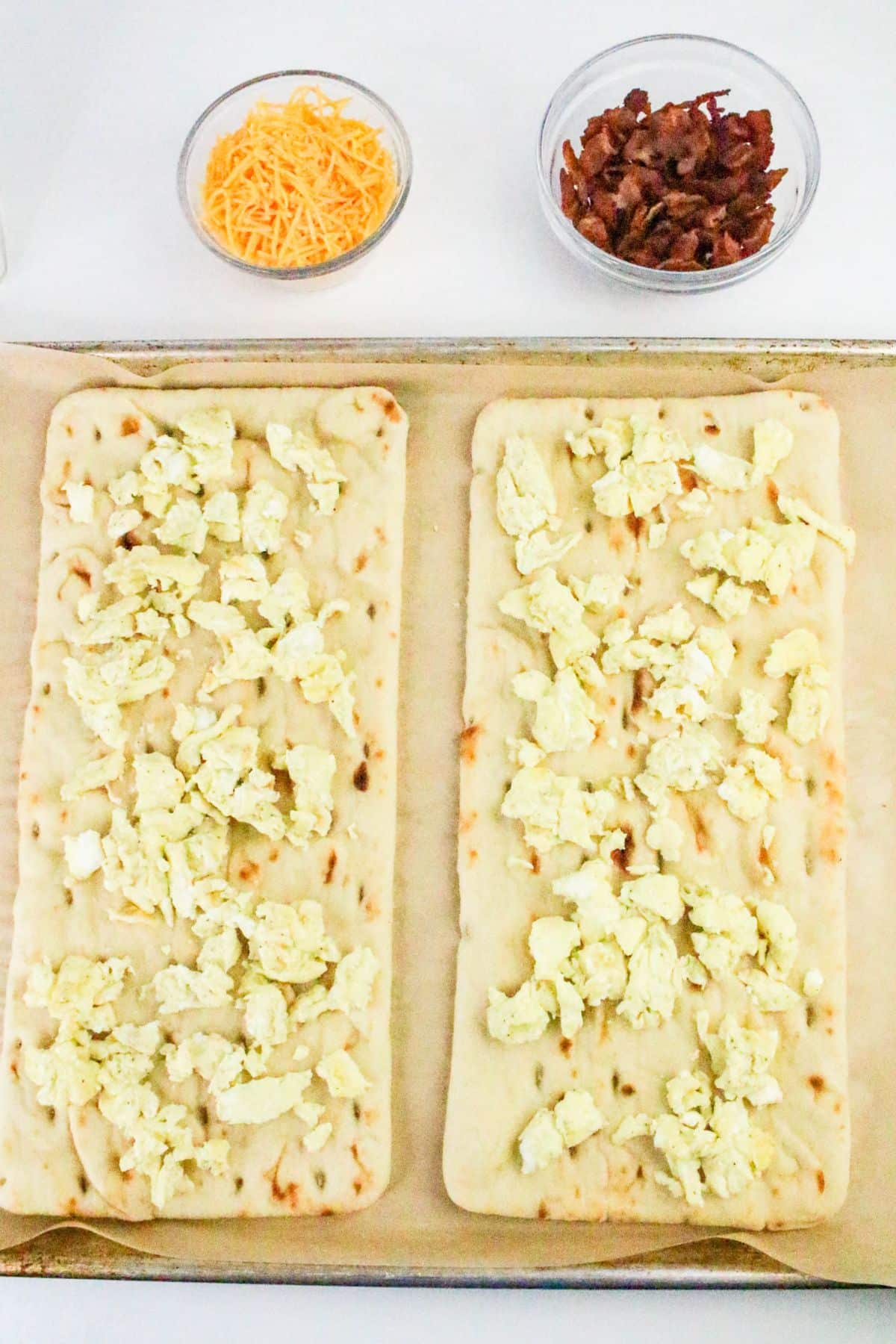 Two rectangular flatbreads topped with scrambled eggs on a baking sheet, with bowls of shredded cheese and cooked bacon pieces above them.