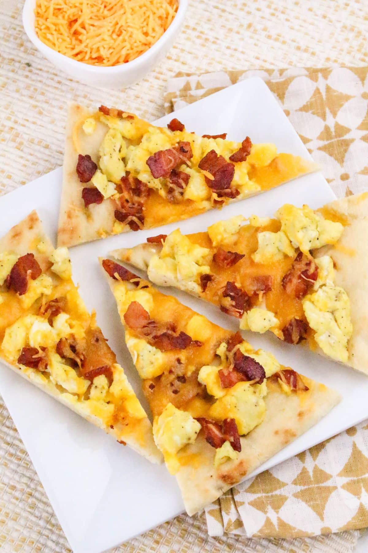 Four slices of breakfast pizza topped with scrambled eggs, bacon, and cheese are arranged on a white plate. A small bowl of shredded cheese is in the background.
