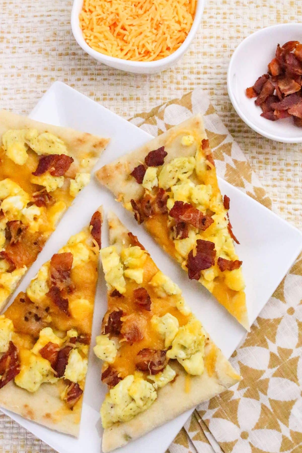 Four slices of breakfast pizza topped with scrambled eggs, cheddar cheese, and bacon on a white plate, with small bowls of shredded cheese and bacon on the side.