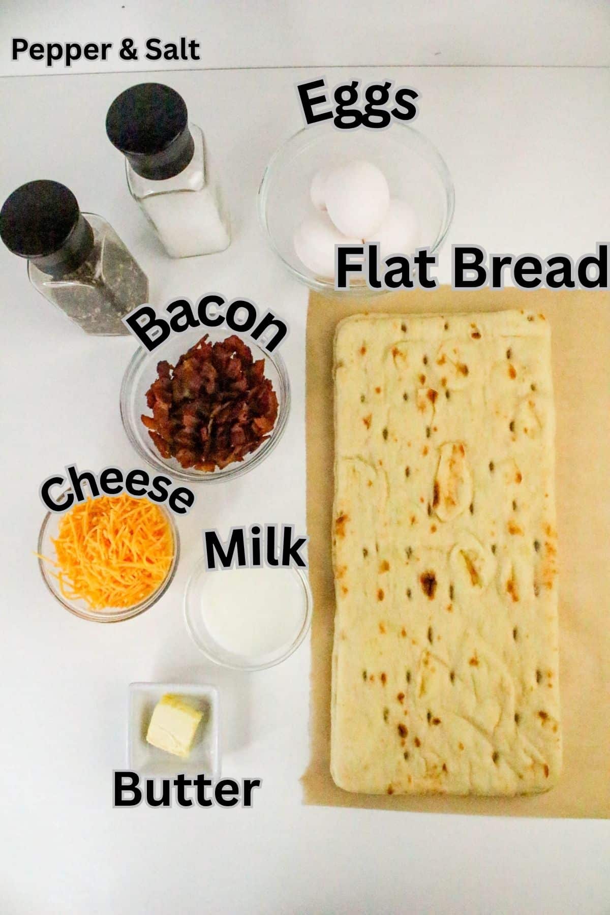 Labeled ingredients for a breakfast recipe are arranged on a white surface, including flat bread, eggs, bacon, cheese, milk, butter, pepper, and salt.