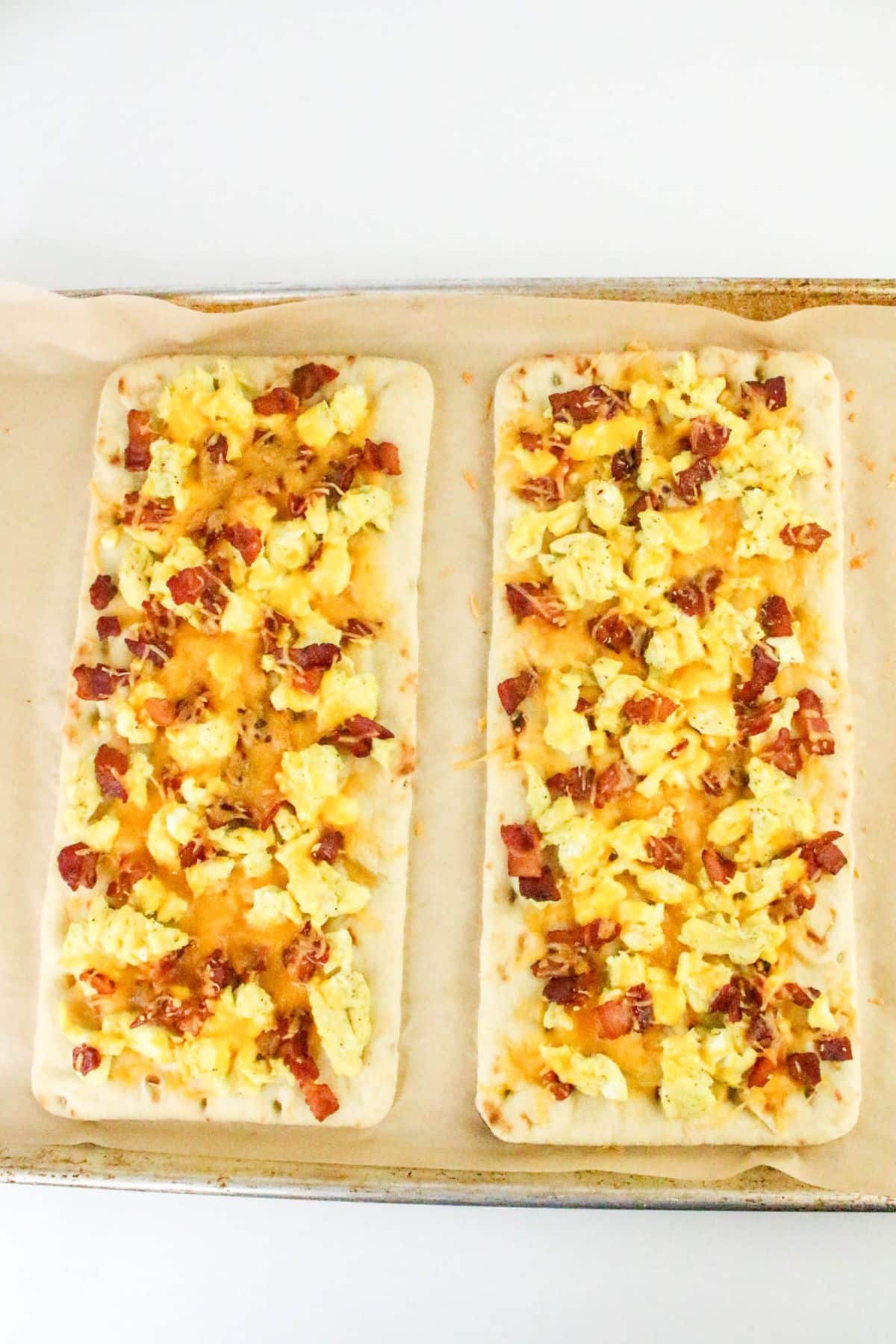 Two rectangular flatbreads topped with scrambled eggs, crumbled bacon, and melted cheese on a parchment-lined baking sheet.