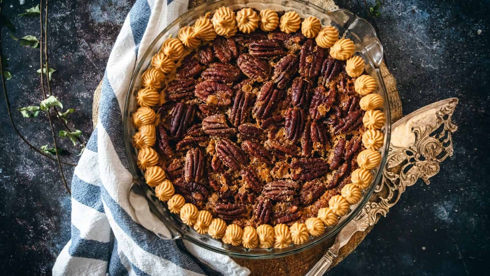 A pecan pie topped with whole pecans and a decorative border of piped cream sits on a glass pie dish next to a striped cloth and ornate pie server.