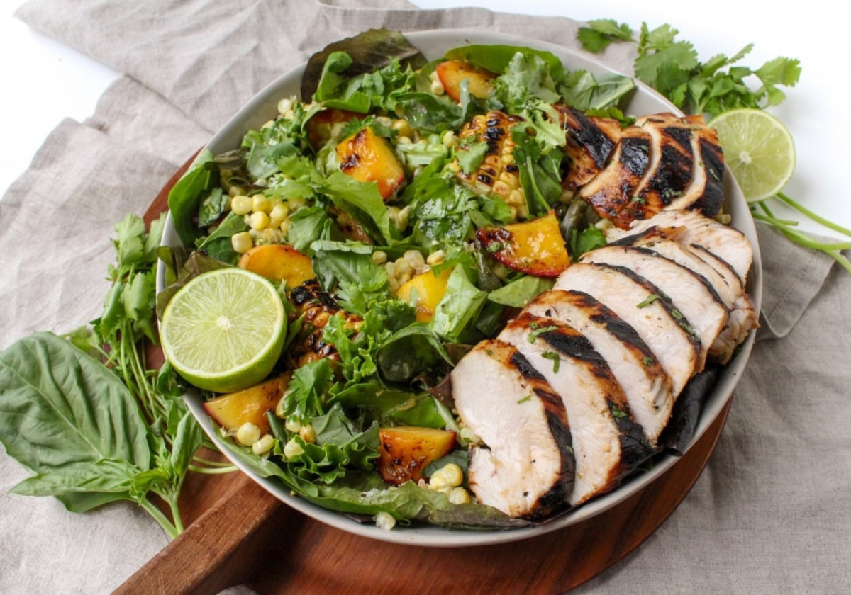 A shallow white bowl with grilled chicken, peaches, corn, and greens with a sliced lime on top.