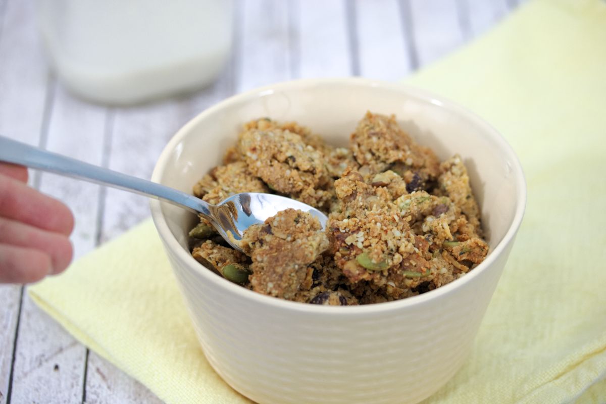 Protein granola in a beige bowl with a hand holding a spoon in it. Bowl on yellow napkin with a milk jug behind it