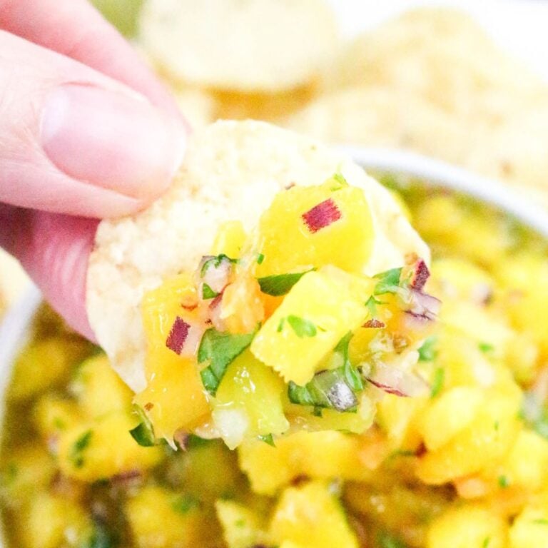 A hand holding a tortilla chip topped with fresh mango salsa containing diced mango, red onion, cilantro, and other ingredients.